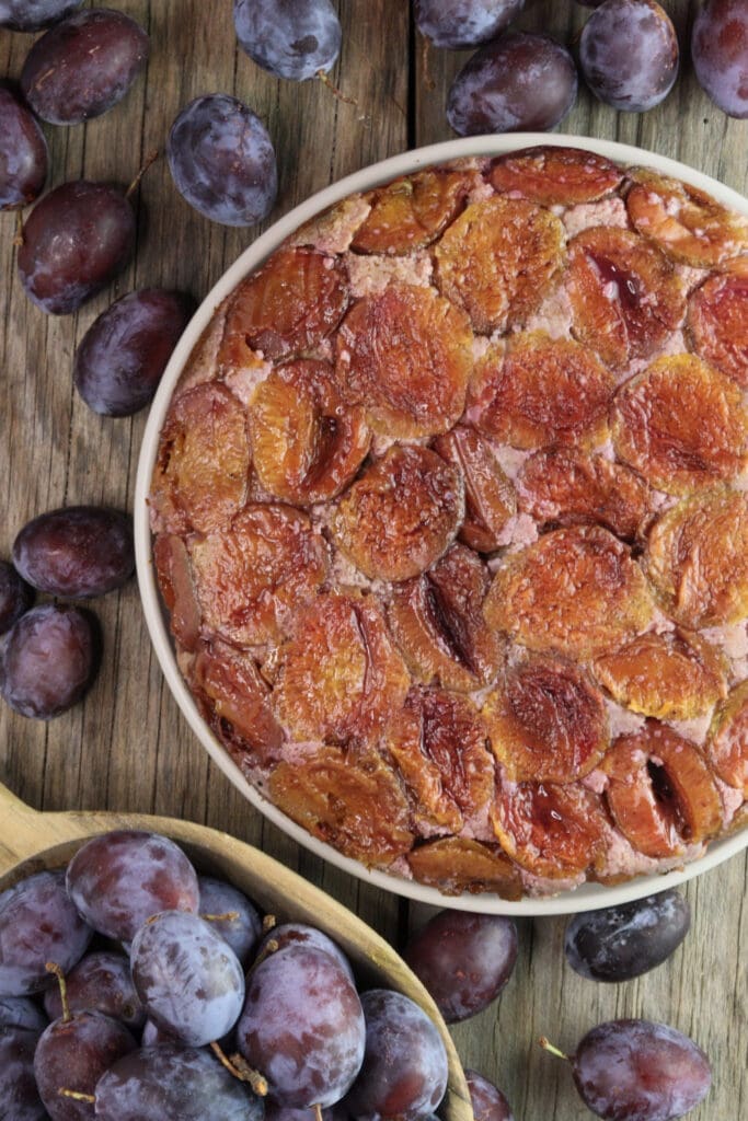 Upside down plum cake with buckwheat on a plate, surrounded with fresh plums on a wooden table